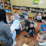 Dzieci siedzą na ziemi i z uwagą słuchają czytanego przez panią opowiadania