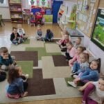 Dzieci siedzą na ziemi i z uwagą słuchają opowiadania