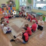 Dzieci siedzą na ziemi i z uwagą słuchają czytanego przez panią opowiadania