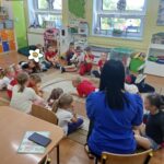Dzieci siedzą na ziemi i z uwagą słuchają czytanego przez panią opowiadania