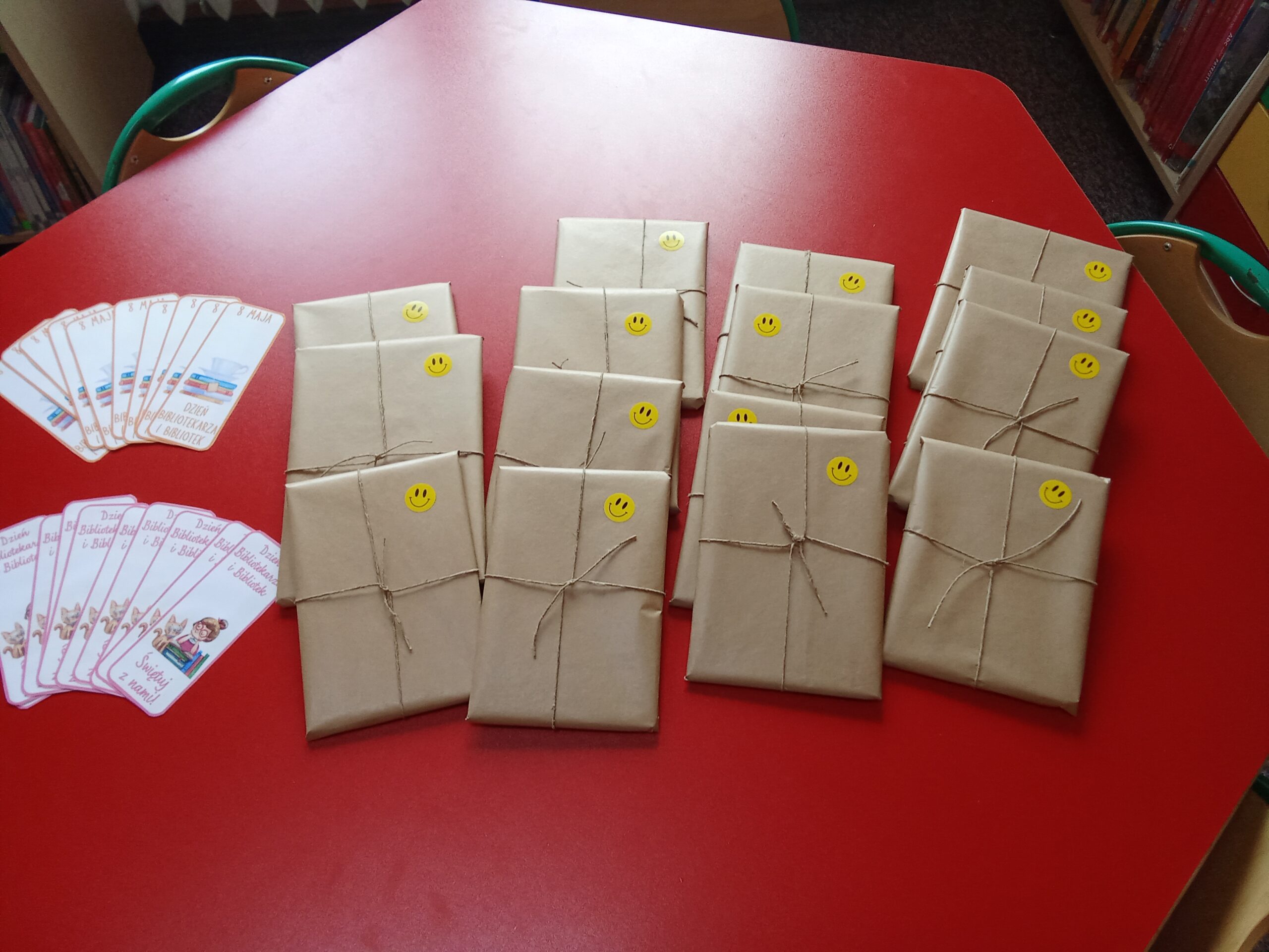 Na czerwonym stole leżą zapakowane w papier książki niespodzianki, obok nich leżą zakładki do książek