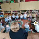 Dzieci siedzą na ziemi i z uwagą słuchają czytanego przez panią opowiadania