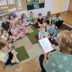 Dzieci siedzą na ziemi i z uwagą słuchają czytanego przez panią opowiadania