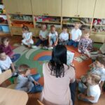 Dzieci siedzą na ziemi i z uwagą słuchają czytanego przez panią opowiadania